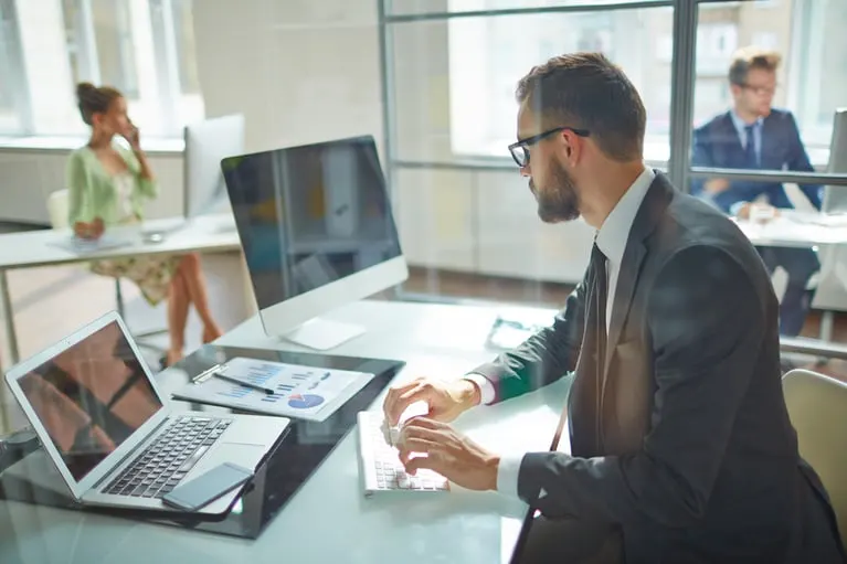 Man working at a standing desk with a monitor and laptop in an office