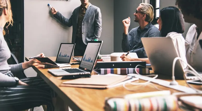 Team meeting with a presenter at a whiteboard and colleagues laughing at a laptop-covered table